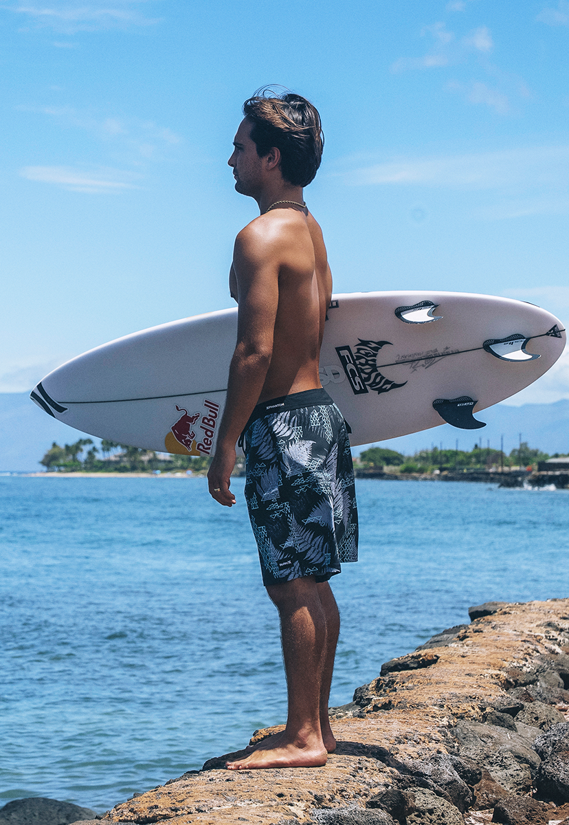 Man holding a surfboard by the ocean with a clear blue sky wearing Hurley boardshorts.