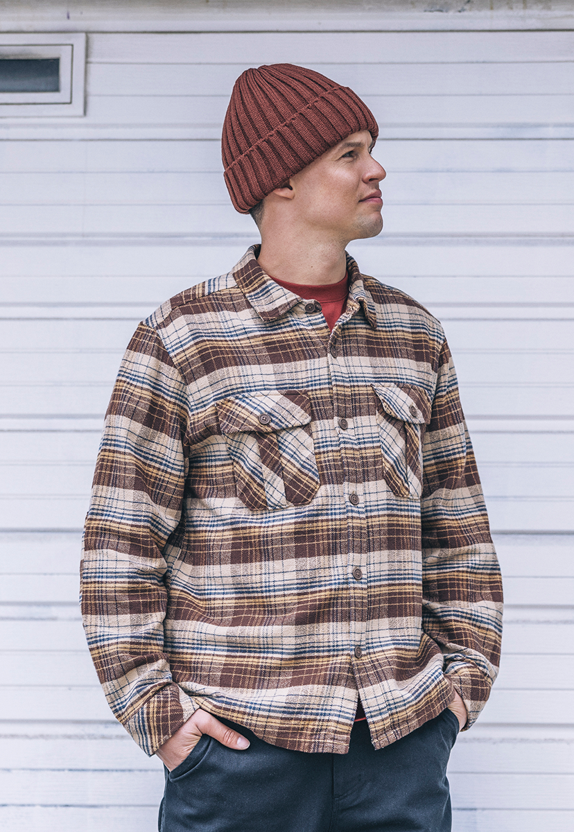 Man wearing a plaid shirt and beanie standing against a white wall.