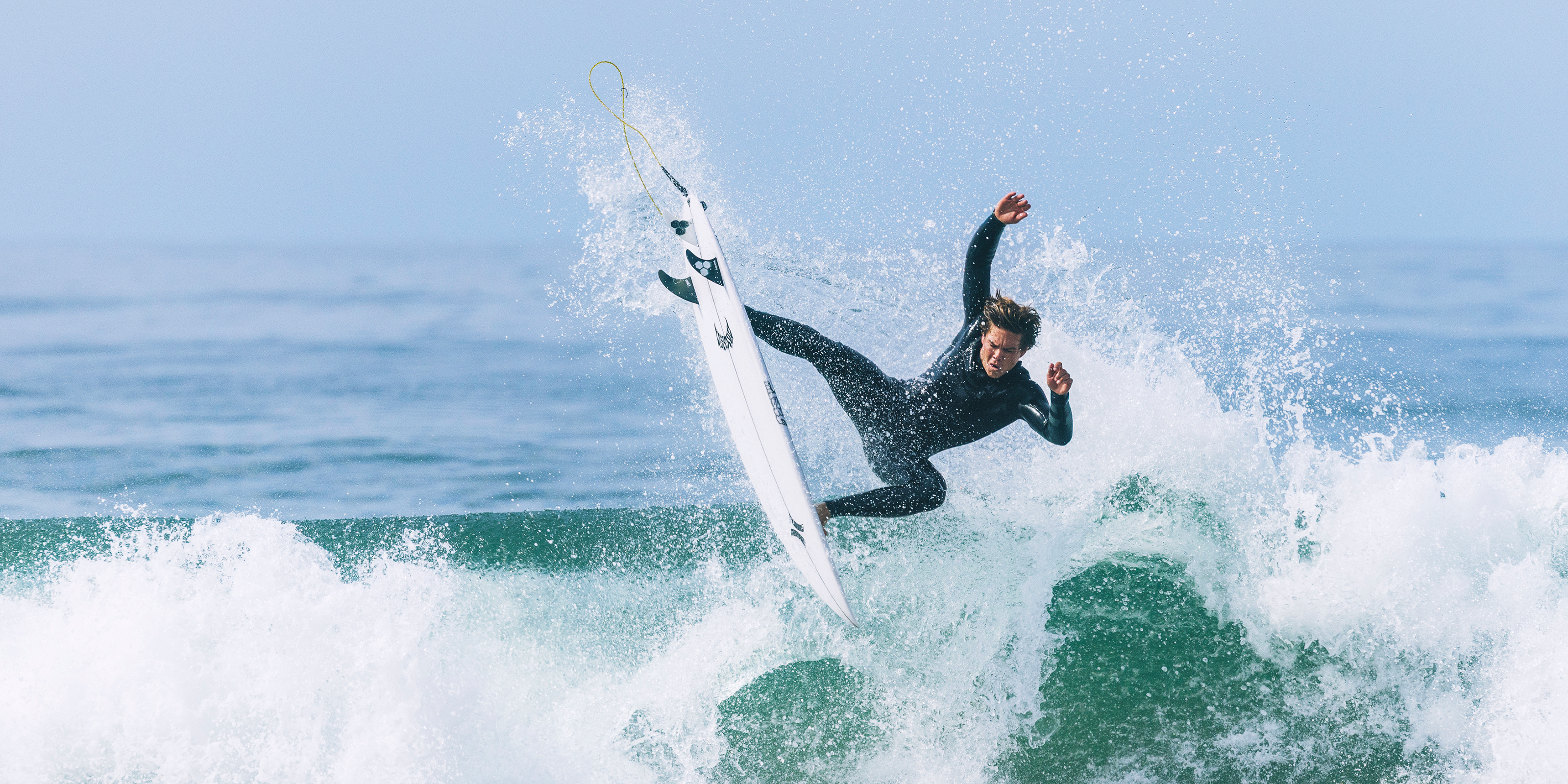 A surfer in a black Hurley wetsuit rides a white surfboard over a breaking ocean wave, with ocean water and spray in the background.