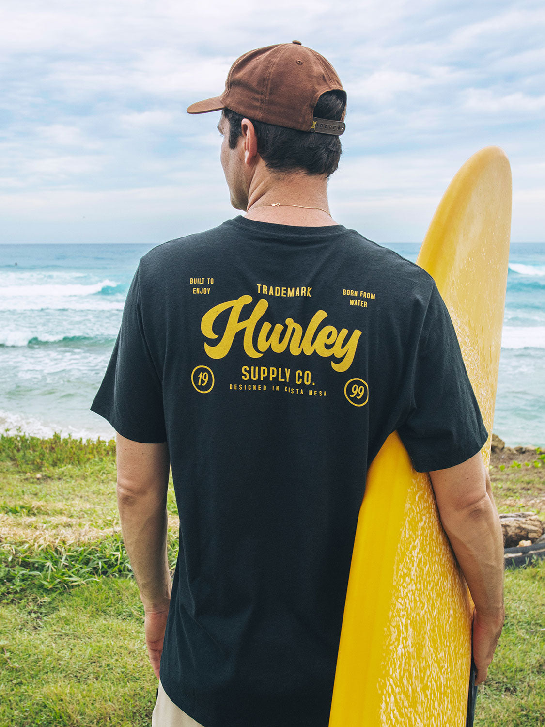 Person wearing a black Hurley t-shirt with yellow text, holding a yellow surfboard by the ocean.